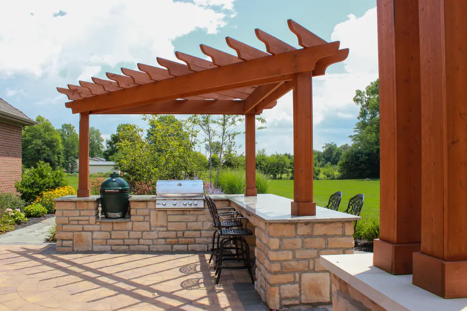 Built-in grill counter under wood pergola with stone base, bar seating, paver patio, and lawn view.